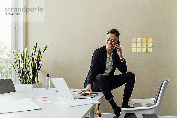 Unternehmerin  die mit dem Handy telefoniert  während sie im Büro einen Laptop benutzt