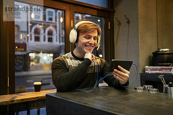 Junger Mann mit Kopfhörern lächelt bei einem Videoanruf auf einem Mobiltelefon in einer Bar