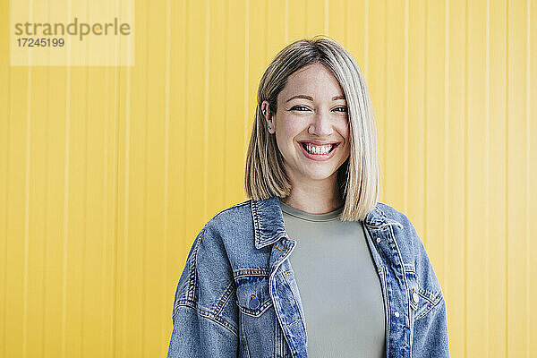 Glückliche blonde Frau lächelnd vor einer gelben Wand