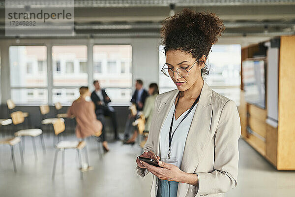 Unternehmerin bei der Nutzung eines Mobiltelefons mit Kollegen im Hintergrund im Büro