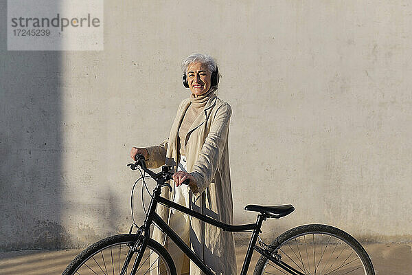 Lächelnde reife Frau mit Kopfhörern  stehend mit Fahrrad