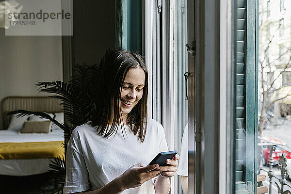 Lächelnde junge Frau benutzt ihr Smartphone am Fenster zu Hause