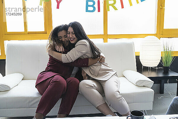 Lächelnde Geschäftsfrauen umarmen sich auf dem Sofa während einer Geburtstagsfeier im Coworking-Büro