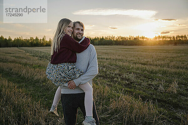 Lächelnder Vater trägt seine Tochter bei Sonnenuntergang auf einem Feld