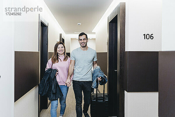 Lächelndes Paar  das sich an den Händen hält  während es mit Gepäck im Hotelflur spazieren geht