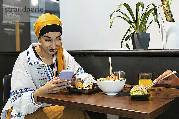 Junge Frau mit Kopftuch benutzt ihr Smartphone  während sie mit einer Freundin im Restaurant sitzt