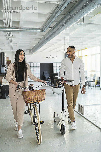 Lächelnder Geschäftsmann  der eine Kollegin ansieht  während er mit Fahrrädern in einem Coworking-Büro spazieren geht