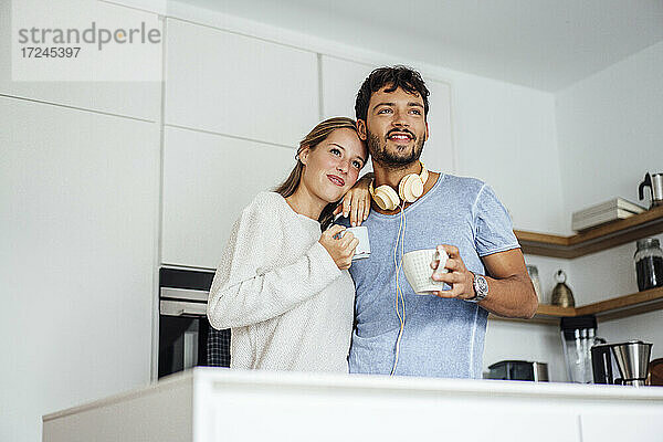 Lächelndes junges Paar hält Kaffeetasse in der Küche und schaut weg