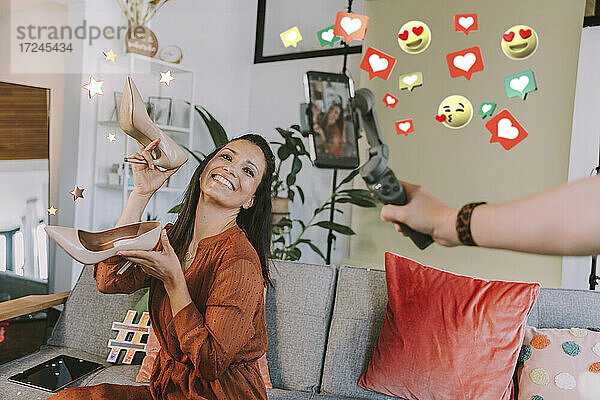 Lächelnde weibliche Influencerin  die High Heels zeigt  während sie von einem Freund mit dem Handy bei sozialen Symbolen zu Hause gefilmt wird