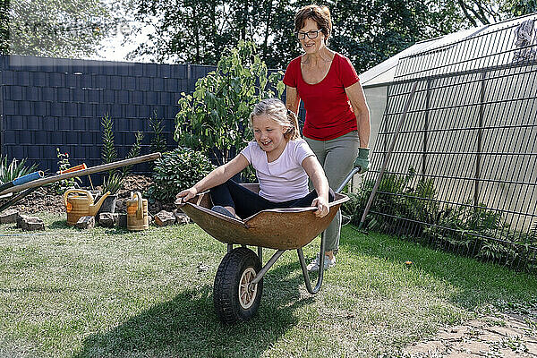 Glückliches Mädchen  das in einer Schubkarre sitzt und mit seiner Großmutter im Hinterhof spielt