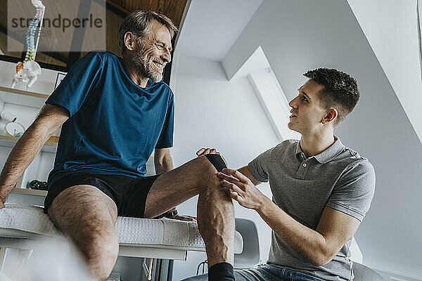 Männlicher Physiotherapeut klebt Kinesio-Tape auf das Knie eines lächelnden Patienten in der Praxis