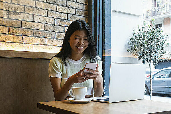 Lächelnde Frau  die ihr Smartphone am Laptop benutzt  während sie in einem Café sitzt