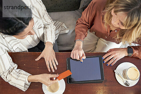 Geschäftsfrau mit Kreditkarte  die mit einer Kollegin zusammensitzt und einen Tablet-PC in einer Cafeteria benutzt