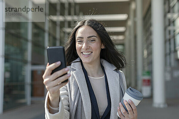 Lächelnde Geschäftsfrau mit Einwegbecher nimmt Selfie durch Smartphone