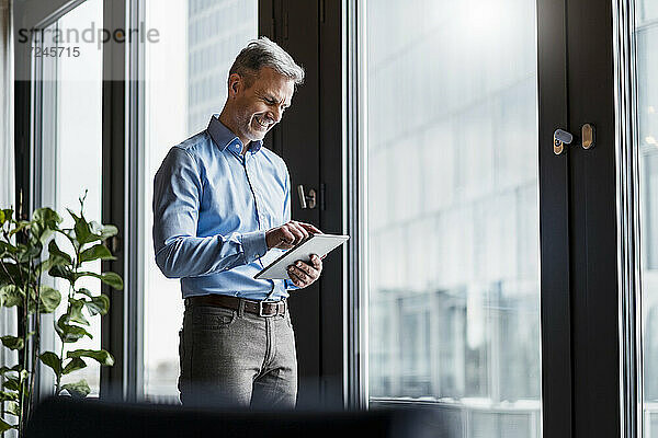 Lächelnder reifer Geschäftsmann bei der Arbeit an einem digitalen Tablet am Fenster am Arbeitsplatz