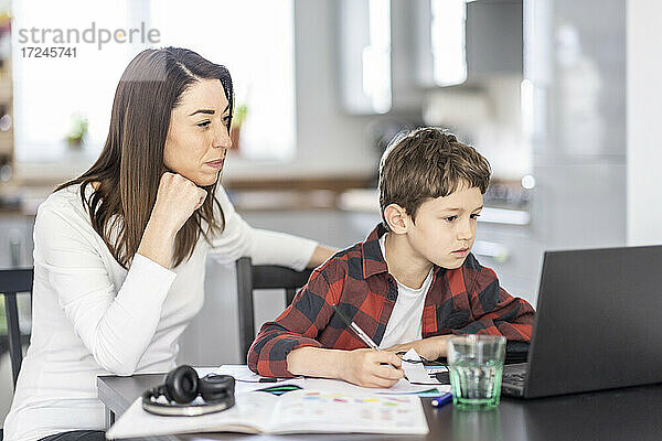 Junge schreibt mit seiner Mutter auf ein Buch  während er zu Hause e-Learning betreibt