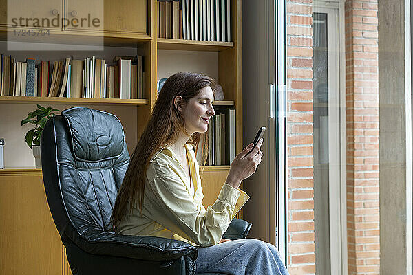 Lächelnde Frau  die ein Mobiltelefon auf einem Stuhl zu Hause benutzt
