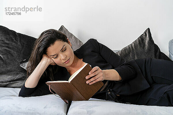 Mittlere erwachsene Frau liest ein Buch und ruht sich zu Hause auf dem Sofa aus