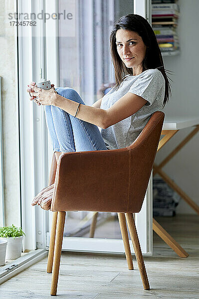 Schöne Frau hält eine Kaffeetasse  während sie auf einem Stuhl zu Hause sitzt
