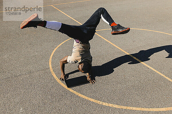 Junger Mann balanciert im Handstand mit einem Grafiktablett auf einem Basketballplatz an einem sonnigen Tag