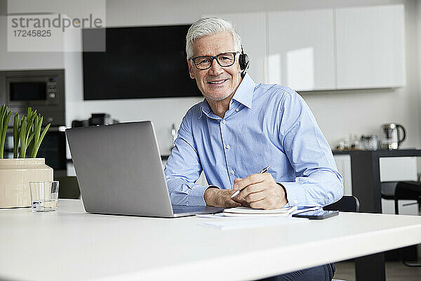 Lächelnder männlicher Unternehmer mit drahtlosem Headset und Laptop am Schreibtisch sitzend