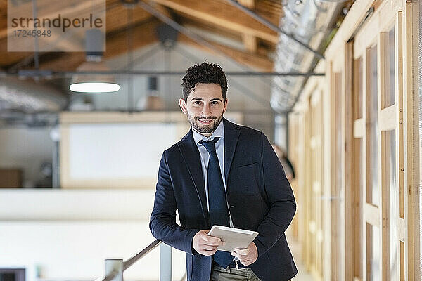 Gutaussehender Mann mit digitalem Tablet  der sich auf ein Geländer im Büroflur stützt