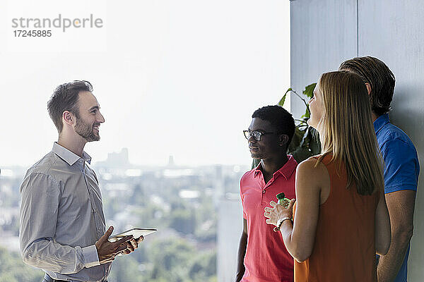 Lächelnde männliche Führungskraft  die ein digitales Tablet hält  während sie mit Kollegen in einer Besprechung im Büro diskutiert