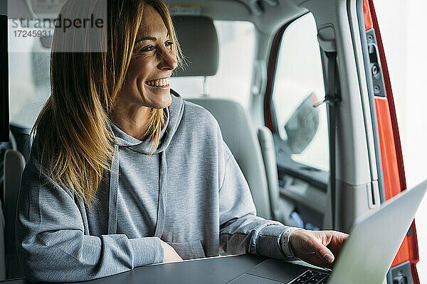 Fröhliche Unternehmerin mit Laptop  die wegschaut  während sie in einem Lieferwagen sitzt