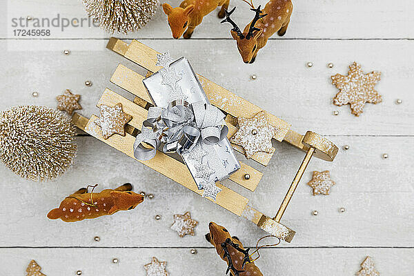 Weihnachtsgeschenk auf Spielzeug-Schlitten  umgeben von Spielzeug-Hirschen mit selbstgebackenen Keksen vor weißem Holzhintergrund