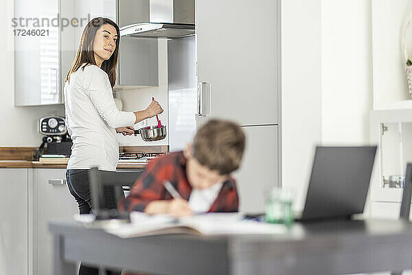 Nachdenkliche Frau bei der Arbeit in der Küche hinter ihrem Sohn  der zu Hause lernt
