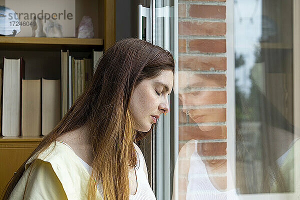 Traurige Frau mit geschlossenen Augen  die sich zu Hause ans Fenster lehnt