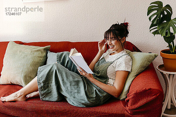 Lächelnde Frau mit Brille liest ein Buch  während sie zu Hause auf dem Sofa sitzt