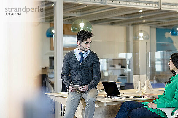 Geschäftsmann mit digitalem Tablet  der eine Kollegin anschaut  während er auf dem Schreibtisch in einem Coworking-Büro sitzt