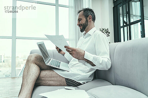 Geschäftsmann mit Laptop  der ein Dokument hält  während er zu Hause auf dem Sofa sitzt