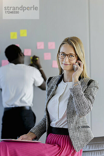 Weibliche Fachkraft  die mit einem Smartphone spricht  während ein männlicher Kollege im Hintergrund im Büro sitzt