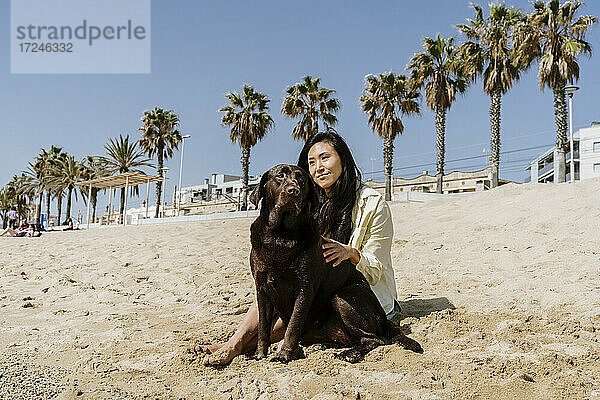 Frau  die ihren Hund streichelt  während sie an einem sonnigen Tag im Sand am Strand sitzt