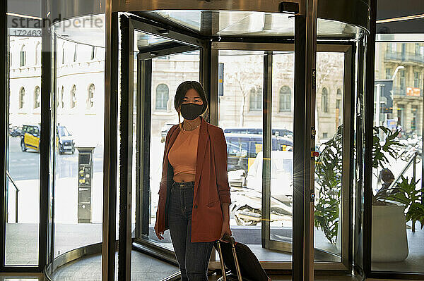 Junge Frau trägt einen Gesichtsschutz  während sie mit ihrem Gepäck am Eingang eines Hotels steht