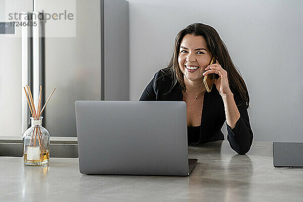 Lächelnde Geschäftsfrau  die mit dem Handy telefoniert  während sie zu Hause am Laptop sitzt