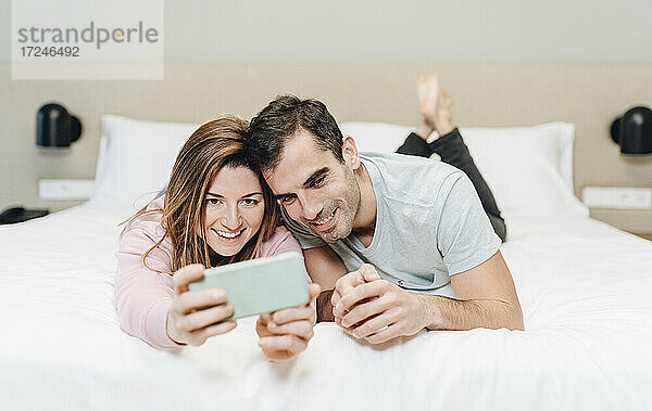 Lächelnde Frau nimmt Selfie mit Mann durch Handy im Hotelzimmer