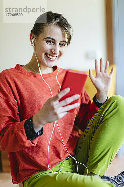 Lächelnde Frau mit In-Ear-Kopfhörern bei einem Videogespräch über das Mobiltelefon zu Hause