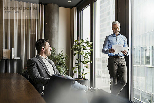 Lächelnder Geschäftsmann  der mit einem männlichen Kollegen über Dokumente am Fenster im Büro diskutiert