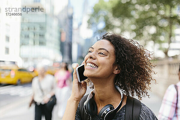 Lächelnde Frau mit Kopfhörern  die in der Stadt mit einem Smartphone telefoniert