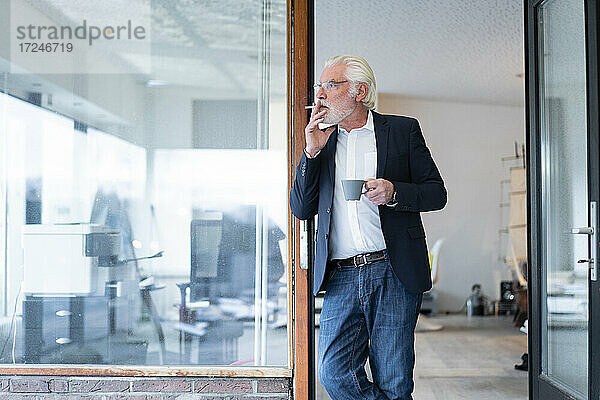 Ein älterer Geschäftsmann raucht eine Zigarette und lehnt sich an eine Glaswand im Büro
