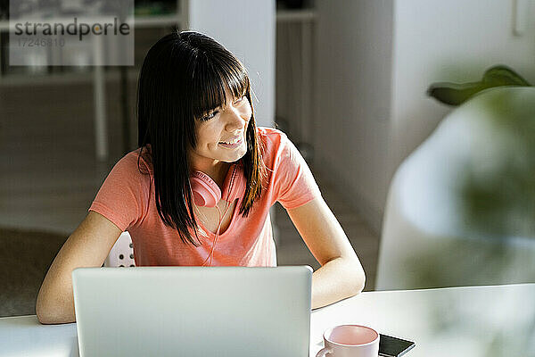 Lächelnde Unternehmerin  die wegschaut  während sie im Büro zu Hause mit einem Laptop sitzt