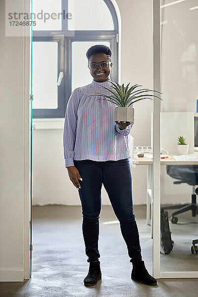 Lächelnde weibliche Fachkraft  die eine Pflanze hält  während sie in der Nähe der Eingangstür im Büro steht