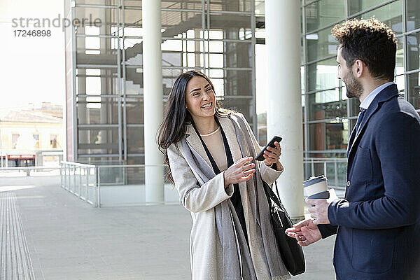 Lächelnde Geschäftsfrau mit Mobiltelefon im Gespräch mit einem Kollegen auf dem Fußweg