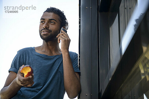 Gut aussehender Mann  der einen Apfel in der Hand hält  während er auf einem Balkon mit seinem Smartphone spricht