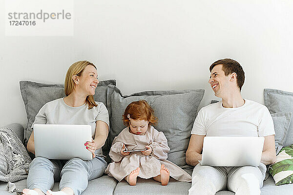 Glückliche männliche und weibliche Unternehmer mit Laptop  die lächelnd neben ihrer Tochter auf dem Sofa zu Hause sitzen