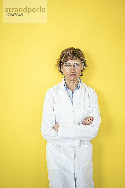 Älterer Wissenschaftler in Arbeitsschutzkleidung lehnt an einer gelben Wand