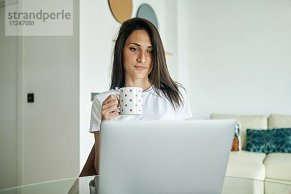 Mittlere erwachsene Frau hält eine Kaffeetasse  während sie zu Hause einen Laptop benutzt